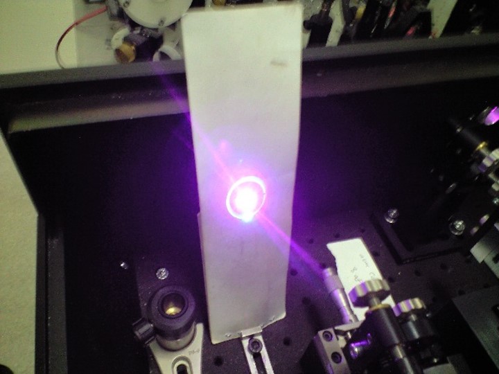 フェムト秒レーザーというとても短い時間に光るレーザーを用いたときの、パラメトリック発振という現象の写真です。中心に虹色の輪ができていて、下の方に緑色の発振光が出ています。10フェムト秒よりも短く光っているもので、様々な光計測に用いられます。光の空間と時間、それと非線形結晶という結晶の角度をうまく調整すると発生します。慣れると簡単に作れますが、熟達するまでは比較的時間がかかります。こういう光技術の組み合わせで最先端の計測技術が成り立っています。