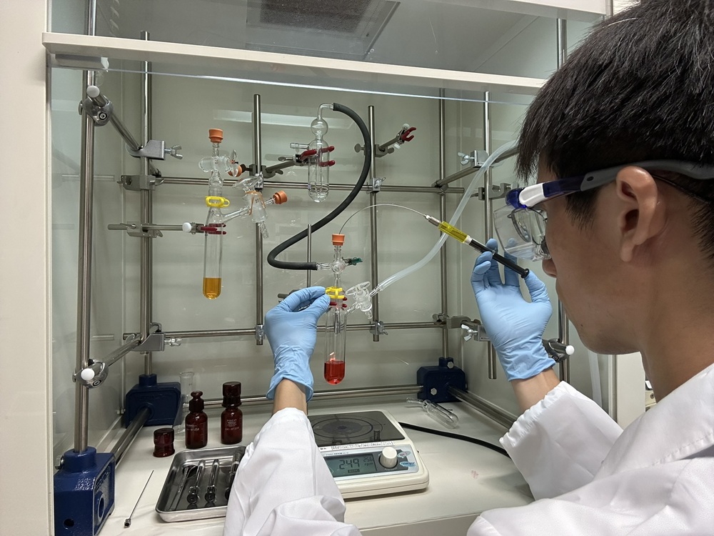化学実験（有機合成）。原料や触媒となる試薬を添加し、特定の条件下で化学反応させることで目的の化合物を合成します。