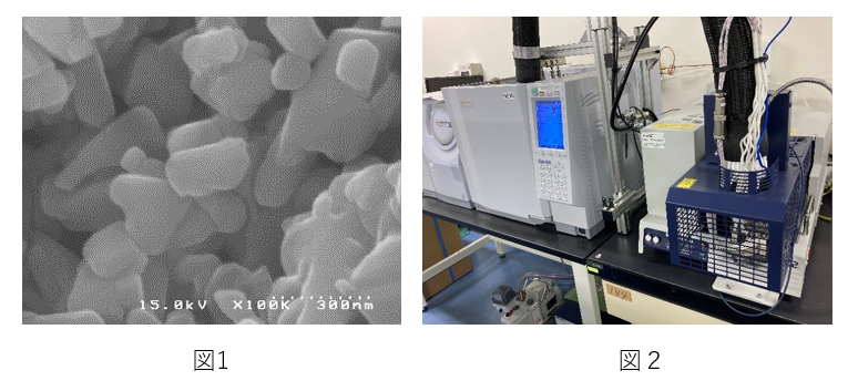 （図１）ナトリウムフェライトの粉末を１０万倍に拡大した電子顕微鏡写真です。300ナノメートル前後のサイズをもつ粒子が観察できます。１ナノメートルは、１０億分の１メートルです。ものすごく小さな領域で二酸化炭素が吸収されます。
（図２）二酸化炭素の吸収量を調べる分析装置です。大学生になると研究室では高度な装置も使えるようになります。