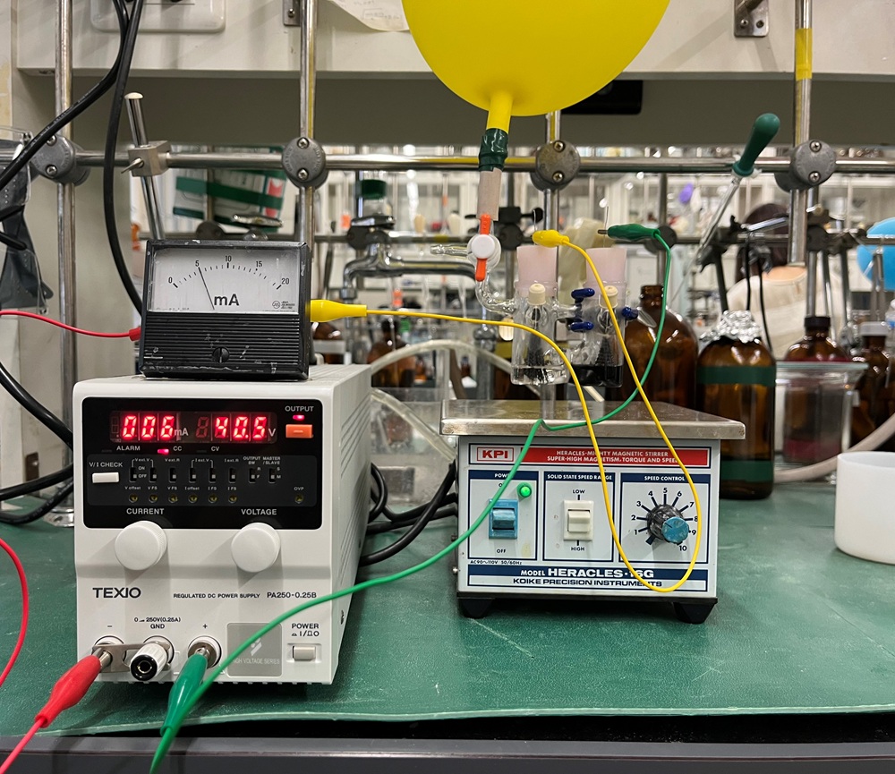 有機電解合成の実験中の写真。H型という真ん中に膜があるガラス器具の両方に電極を差し込み、直流の電気を流して、有機化合物の分子変換を行うことができます。