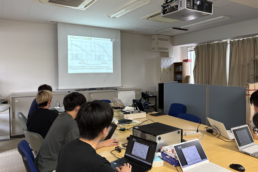 研究室のゼミの様子です。参加者はノートパソコンで資料を閲覧し、Zoomを用いたハイブリッド形式で学外の方も参加しています。