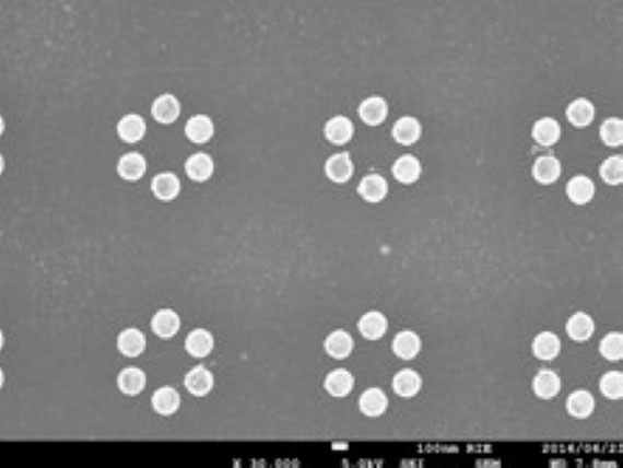 電子顕微鏡写真。製作した金属ナノ粒子の一例です。写真中の円形に写っている物体が金のナノ粒子でその大きさは100 nm（1nmは 1/10,000,000cm）くらいです。六角形状に並んでいて、ベンゼンのように見えないでしょうか。実際個々の金ナノ粒子を“原子”、６個並べると“分子”と見立てると、原子と分子を繋ぐ共有結合に類似した効果が見られます。